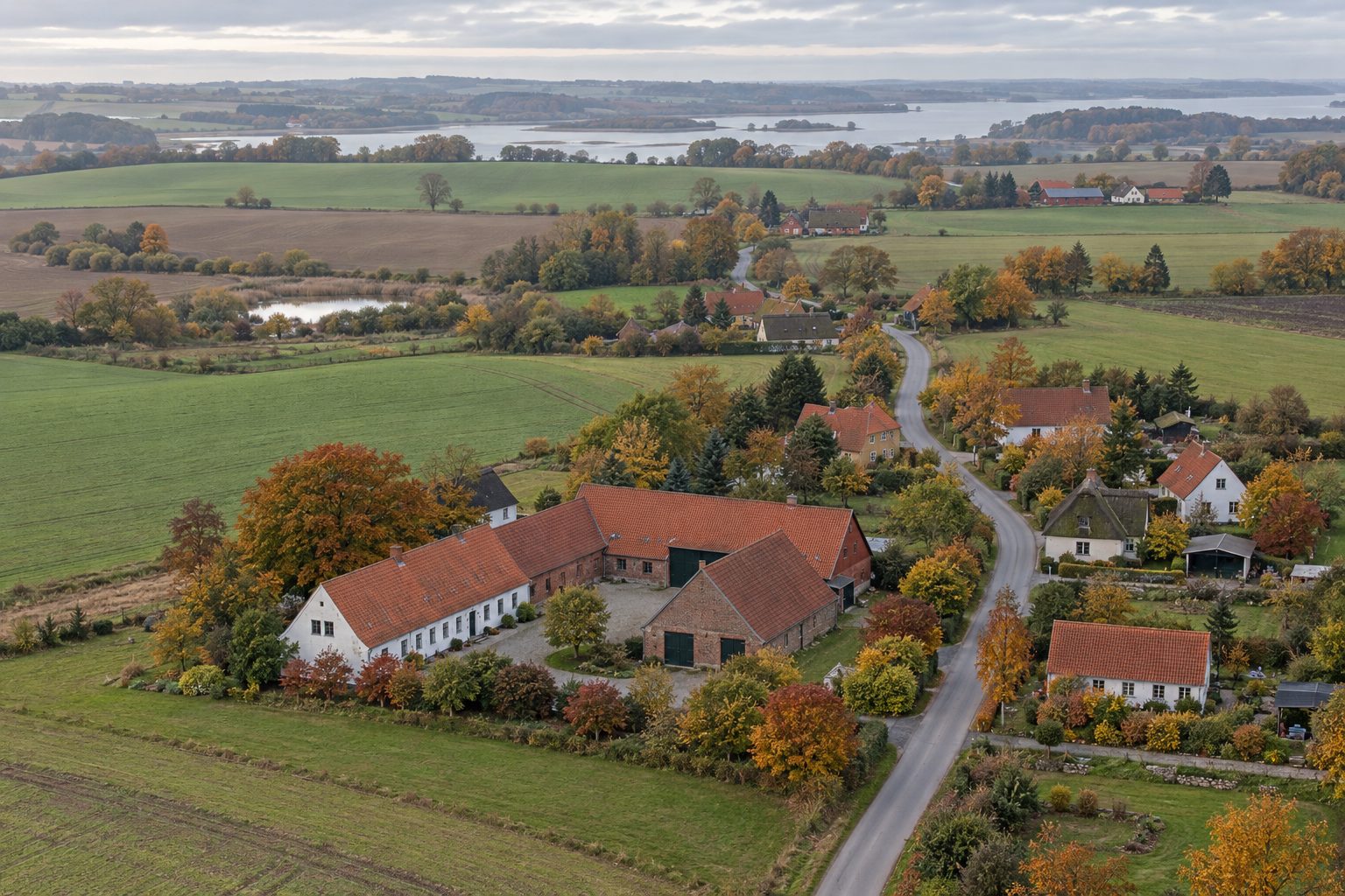 Et tænkt dansk landsbymiljø set skråt fra luften. AI-genereret temabillede; ikke et historisk arkivfoto eller en bestemt virkelig lokalitet.