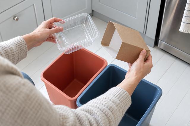 Hænder adskiller pap og plast ved to affaldsbeholdere. AI-genereret temabillede; farverne er ikke en konkret sorteringsvejledning.